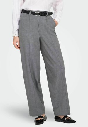 Femme portant un pantalon large taille haute gris avec une ceinture noire, une blouse blanche et des chaussures plates noires à clous, debout les mains dans les poches.