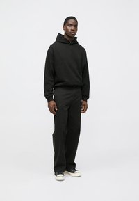 Čierna mikina s kapucňou a voľným strihom, doplnená voľnými čiernymi nohavicami a bielymi teniskami. Materiál pôsobí mäkko, s minimálnymi detailmi.