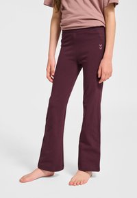 Dziecko ubrane w purpurowe rozszerzane spodnie i kurzawo-różową koszulkę z krótkim rękawem, stojące boso na jednolitym białym tle.