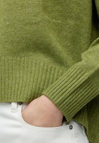 Main partiellement dans la poche d'un pantalon blanc, portant un pull vert en maille avec poignets et ourlet côtelés.