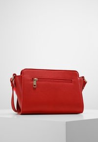 Czerwona, syntetyczna torba na ramię o prostokątnym kształcie, wyposażona w przednią kieszeń na zamek oraz złote elementy ozdobne. Gładka faktura i regulowany pasek.