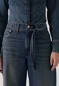 Un jean en denim foncé présente une ceinture avec un nœud, des accents cousus et des poches standard. La texture est lisse avec une légère brillance.