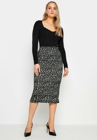 Musta ribbed pitkähihainen paita yhdistettynä tiukkaan mustaan hameeseen, jossa on abstrakti valkoisen ja vihreän täplikkäin kuvio, ja asustettu mustilla korkokenkillä.