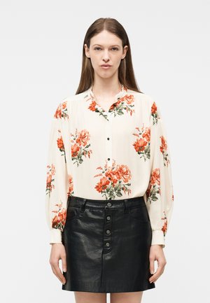 Γυναίκα που φοράει κρεμ μπλούζα με κόκκινα-πορτοκαλί λουλούδια και μαύρη δερμάτινη φούστα με κουμπιά, στέκεται μπροστά σε ανοιχτό γκρι φόντο.