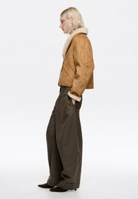 Chaqueta corta de ante marrón con un cuello de borreguito crema, combinada con pantalones anchos verde oscuro y tacones marrones puntiagudos.