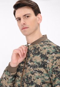 Camouflage bomberjack in tinten van groen, bruin en zwart met een gebreide kraag en volle rits, met een digitaal patroon.