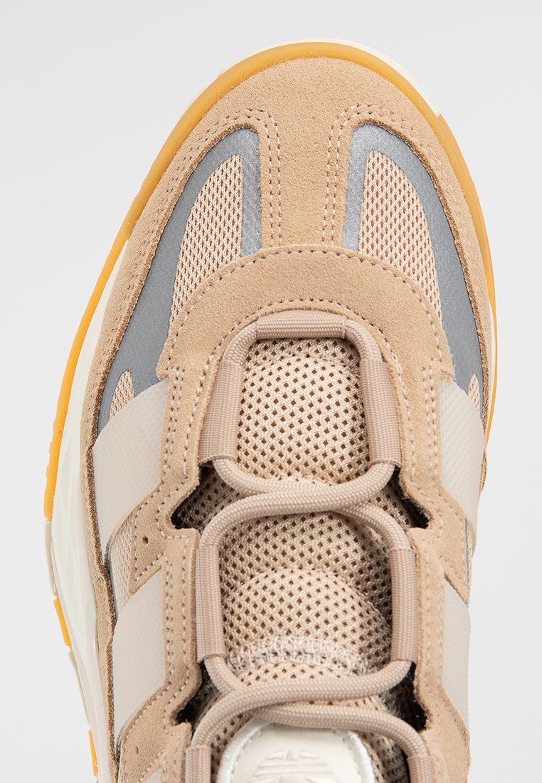 Beige Sportschuh mit Obermaterial aus Mesh und Wildleder, grauen Akzenten und einer strukturierten Gummisohle. Besitzt drei kontrastierende weiße Streifen.