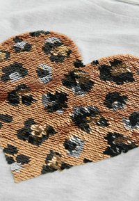 Ukras u obliku srca s korištenjem šljokica u brončanoj, crnoj i srebrnoj boji na svijetlosivoj tkanini, s dizajnom u leopard uzorku i teksturiranom površinom.