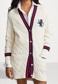 Vit kabelstickad cardigan med djup V-hals, röd och marinblå randig kant och logotypbroderi. Inkluderar framfickor och knappar.