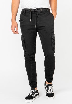 Mann trägt schwarze Cargohosen mit Tunnelzugbund und schwarze Sneaker mit weißen Schnürsenkeln, steht vor weißem Hintergrund.