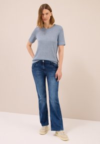 Frau steht da und trägt ein hellblaues Kurzarmoberteil, dunkelblaue Jeans und beige Sneakers vor einem einfarbigen beige Hintergrund.