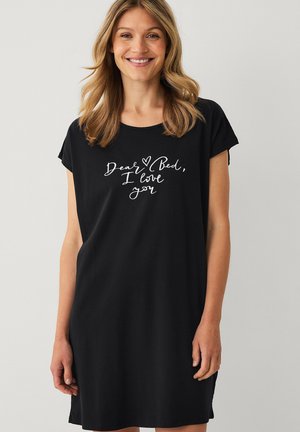 Lachende vrouw met blond haar draagt een zwarte jurk met witte tekst ""Dear Bed, I love you"" op de voorkant tegen een lichte achtergrond.