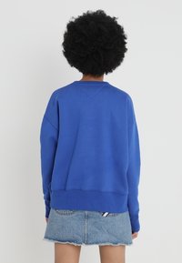 Blue cotton sweatshirt with dropped shoulders and ribbed cuffs, paired with a denim skirt. Simple design, no visible patterns or hardware.