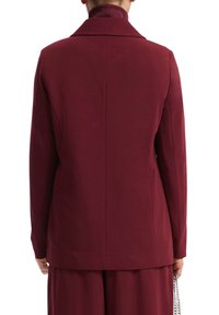 Blazer bordeaux con una texture liscia, silhouette strutturata, rever a sceli, design senza bottoni e cuciture pulite sul retro.