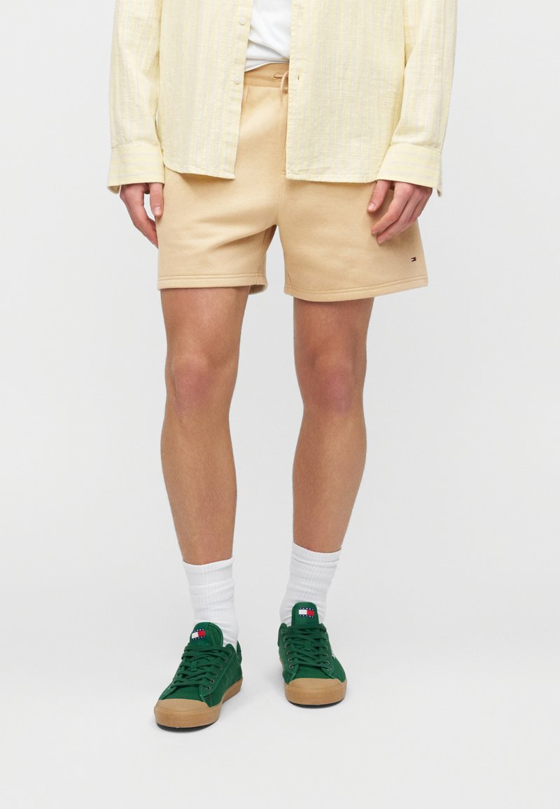 Καφέ βαμβακερά σορτς με ελαστική μέση, συνδυασμένα με ανοιχτό κίτρινο πουκάμισο με κουμπιά και πράσινα αθλητικά παπούτσια με λαστιχένια σόλα. Ορατές λευκές κάλτσες.