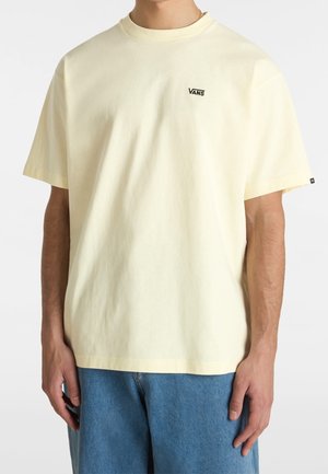 Osoba ubrana w bladożółtą koszulkę Vans i niebieskie dżinsy, stojąca na tle białego tła, z rękami swobodnie opuszczonymi wzdłuż ciała.