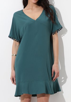 Donna che indossa un abito largo color verde petrolio con scollo a V, maniche corte decorate con pon pon e un orlo arricciato, in piedi contro un muro bianco.