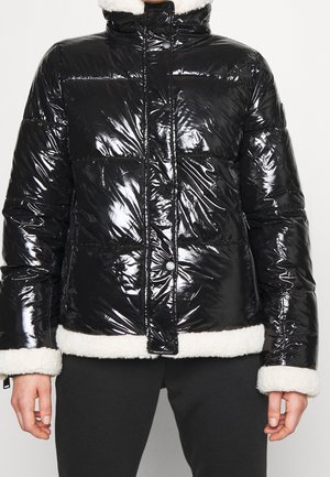 Doudoune noire brillante avec bordure en polaire blanche aux poignets et à l'ourlet, fermeture éclair et boutons-pression, portée sur un pantalon noir.