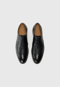 Une paire de chaussures de ville en cuir brogue noir avec des détails texturés et des lacets, vue de dessus sur un fond blanc.