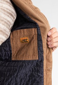 Brown jacket with a quilted navy lining. Includes a brown corduroy patch pocket displaying a multicolored label. Soft texture.