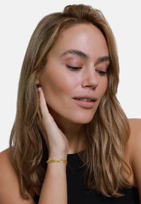 Bracciale in oro su un polso, composto da forme ovali collegate. Capelli lunghi e lisci di colore castano, indossa un top senza maniche di colore nero. La pelle appare liscia.