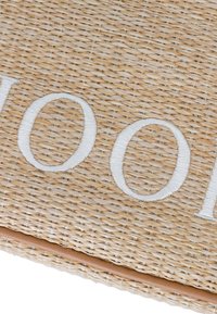 Dettaglio ravvicinato di un tessuto di paglia intrecciato beige con lettere bianche ricamate e una rifinitura in pelle tan lungo il bordo inferiore.