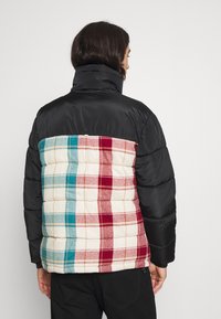 Chaqueta de plumas con un frontal negro y un reverso a cuadros en rojo, turquesa y crema. La parte trasera presenta una textura acolchada y un diseño de bloques de color.