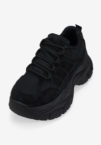 Zapatilla deportiva negra que presenta una combinación de materiales de ante texturizado y malla, punta redondeada, suela gruesa y diseño de cordones.
