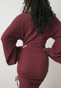 Maroon textuur jurk met lange belmouwen en een aansluitende silhouet. Heeft een geraffineerde taille met trekkoordaccenten voor aanpassing.
