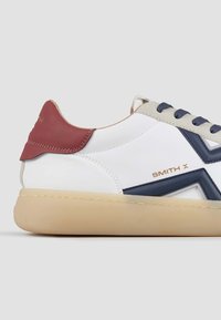 Baskets en cuir blanc avec un design rouge et marine, dotées d'une semelle texturée, de lacets beiges et d'un détail de logo métallique sur le côté.