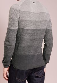 Homme portant un pull en tricot ombré gris avec des manches longues et un jean en denim foncé, vu de dos contre un fond uni.