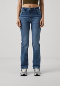 Blå denimjeans med høyt liv og rett passform, med klassisk fem-lommer-design og kontrastfarget søm. Brukes med sølvfargede joggesko.