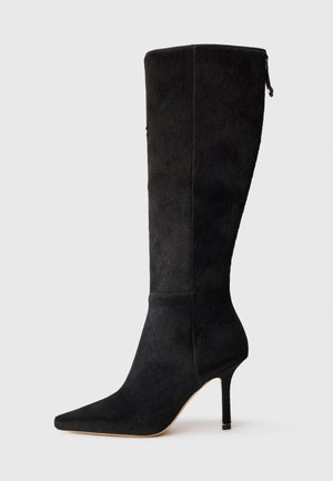 Μαύρη γόβα με ψηλό τακούνι, φτιαγμένη από υλικό με υφή, διαθέτοντας μυτερή μύτη και κομψό σχέδιο που εκτείνεται μέχρι το μέσο της γάμπας, με κλείσιμο τύπου φερμουάρ.