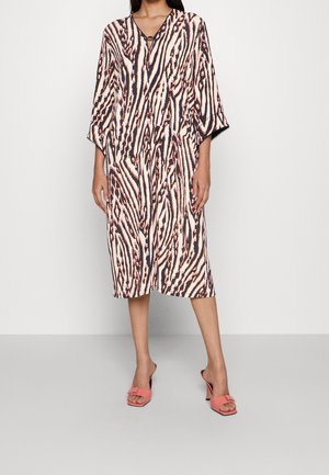 Femme portant une robe mi-longue à rayures zèbre noires, blanches et pêche et des sandales à talons ouvertes corail sur un fond uni.