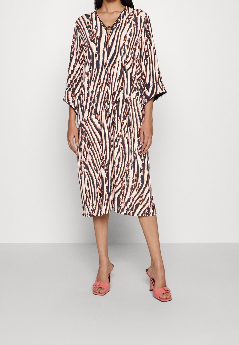 Femme portant une robe mi-longue à rayures zèbre noires, blanches et pêche et des sandales à talons ouvertes corail sur un fond uni.