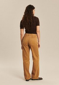 Pantalons larges en velours côtelé couleur brun doré. Comprend des côtes verticales, des poches arrière et une taille haute. Associés à un haut ajusté de couleur sombre.