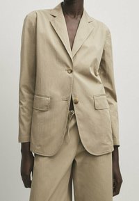 Beżowa bawełniana marynarka z klapami z wycięciem, dwoma przednimi kieszeniami i zapięciem na dwa guziki, w zestawie z beżowymi spodniami. Gładka faktura.