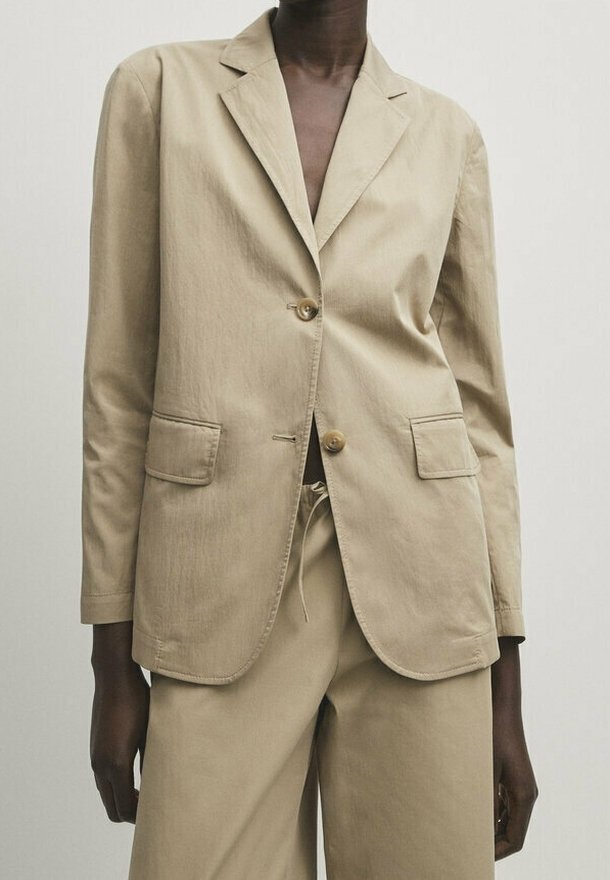 Beżowa bawełniana marynarka z klapami z wycięciem, dwoma przednimi kieszeniami i zapięciem na dwa guziki, w zestawie z beżowymi spodniami. Gładka faktura.