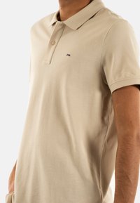 Béžové polo tričko vyrobené z bavlny, vybavené dvouknoflíkovou légou a malým vyšitým logem na hrudi. Krátké rukávy a texturovaná látka.
