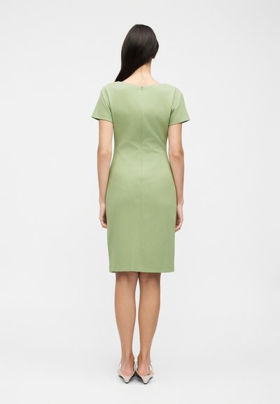 Γυναίκα με μακριά σκούρα μαλλιά που φοράει ένα εφαρμοστό ανοιχτοπράσινο φόρεμα μέχρι το γόνατο με κοντά μανίκια, όρθια πλάτη στην κάμερα σε λευκό φόντο.