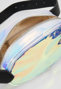 Holograficzna saszetka biodrowa o gładkiej, błyszczącej powierzchni, czarnym pasku, srebrnym suwak oraz beżowej kieszeni wewnętrznej na suwak.