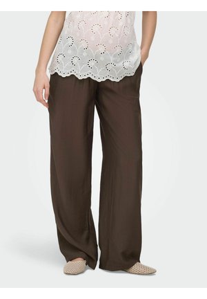 Pantalon ample marron foncé porté avec un top en dentelle ajourée blanche et des chaussures plates tissées beiges, modèle debout avec une main dans la poche.