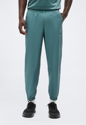 Mees, kes kannab türkiissiniseid dressipükse elastsete mansettidega, käed taskutes, valged sokid ja mustad spordijalatsid, seisab valge tausta ees.