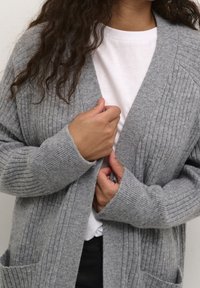 Gray ribbed cardigan with long sleeves, open front, and side pockets, layered over a white t-shirt, showcasing a soft texture.