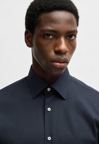 Camicia a maniche lunghe blu navy realizzata in tessuto liscio, con colletto a punta e cinque bottoni bianchi sul davanti.