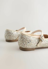 Beez sparkling Mary Jane flats kuldse ümmarguse klambriga heledal neutraalsel pinnal ja taustal.