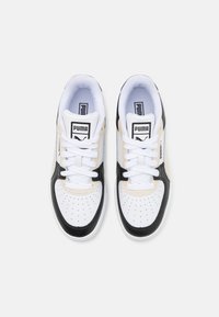 Tenisky Puma s bílou koženou základnou, černými akcenty a béžovými panely. Obsahují perforovanou špičku, ploché bílé tkaničky a štítky s logem.