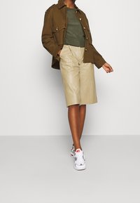 Olijfgroene geribde gebreide trui, bruine canvas jas met knoopdetails, crèmekleurige neplederen shorts en witte sneakers met roze accenten.