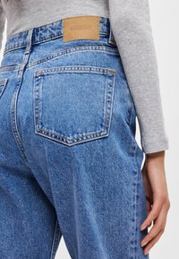 Jasnoniebieskie dżinsy o luźnym kroju, z brązowym skórzanym logo na pasku i dwiema tylniymi kieszeniami z przeszytymi detalami.