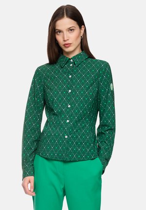 Woman wearing a green button-up shirt with a white diamond and heart pattern, paired with solid green pants against a light background.